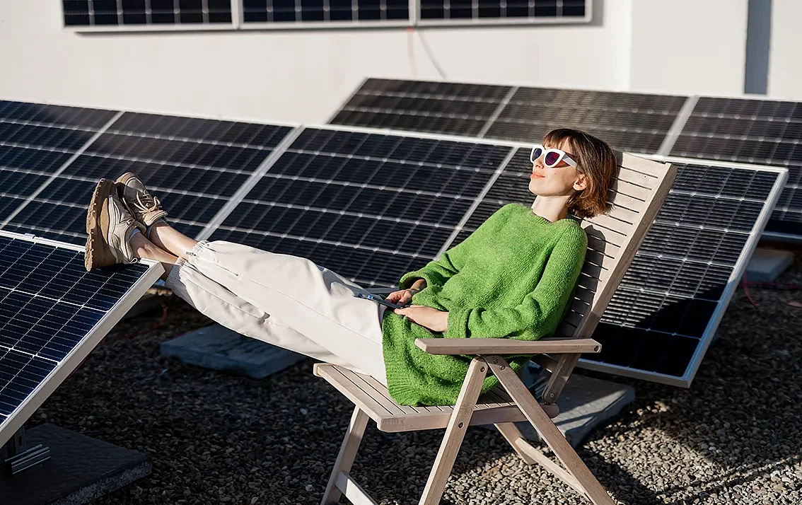 eine Frau sitzt in einem Liegestuhl vor mehreren Solarmodulen auf einem Dach und legt die Füße auf einem Modul hoch
