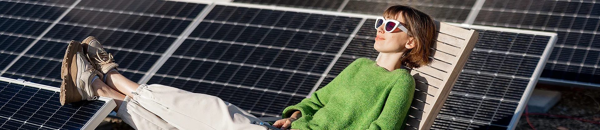 Eine Frau liegt mit Sonnenbrilel in einem Liegestuhl und hat die Füße auf Solarmodule gelegt, die auf dem Flachdach neben ihr aufgebaut sind