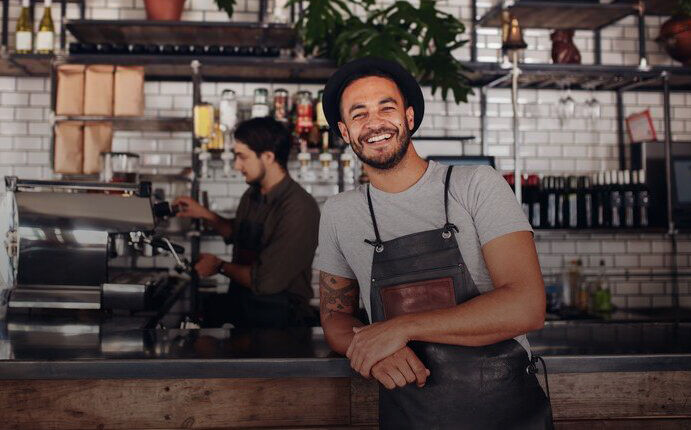 Mann mit einem Hut steht angelehnt, lachend an eine Kaffeebar, im Hintergrund steht eine Person an der Kasse