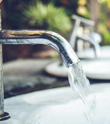 Wasserhahn Wasserhahn mit fließendem Wasser im Vordergrund, während im Hintergrund mehrere Wasserhähne und eine grüne Landschaft zu sehen sind