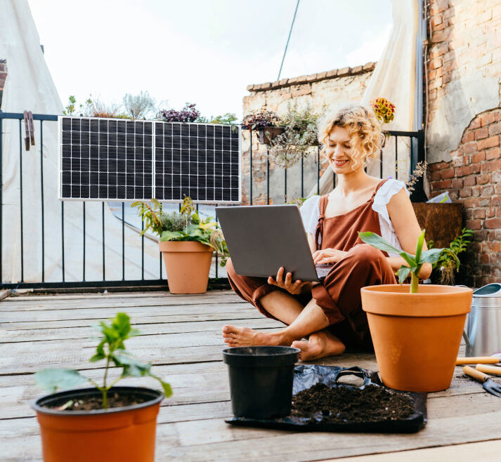 Frau sitzt auf Balkon mit Mini-Solaranlage und ein Laptop 