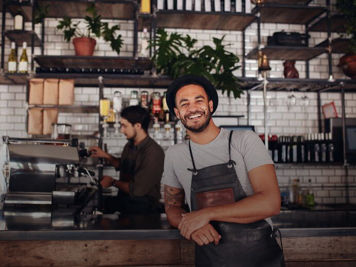 Mann mit einem Hut steht angelehnt, lachend an eine Kaffeebar, im Hintergrund steht eine Person an der Kasse