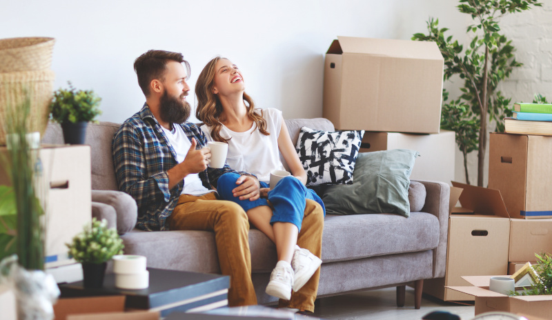 Junges Pärchen glücklich nach Umzug Junges Paar, das lachend auf einem Sofa sitzt, umgeben von Umzugskisten.