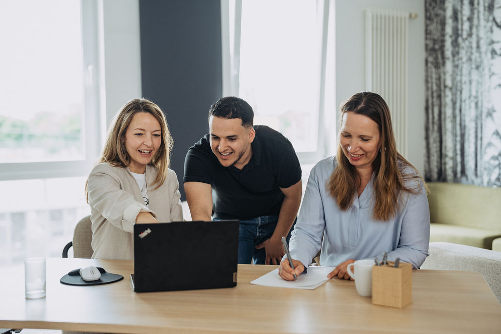zwei Personen schauen auf einen Laptop und eine Frau schriebt auf einen Block
