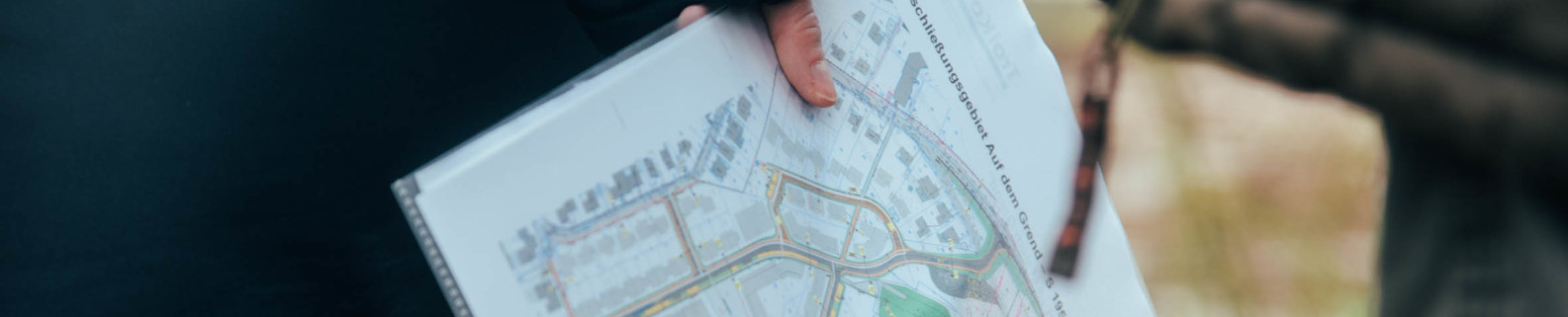 Person hält einen Bauplan in der Hand auf dem Straßen und Gebäude zu sehen sind 